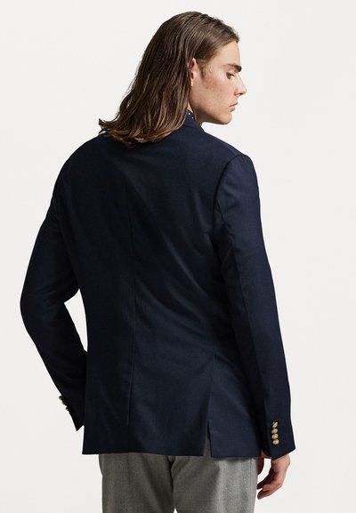 Blazer ajustado azul-marinho com costas ventiladas, textura suave e detalhes em botão dourado nas mangas. Estilizado com calças cinzentas.