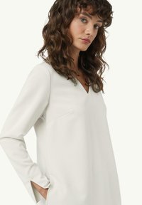 Weißes langärmliges Kleid mit V-Ausschnitt, aus glattem Stoff gefertigt. Verfügt über seitliche Taschen und dezente Nahtdetails. Figurbetonte Passform.