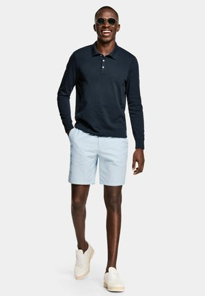 Man glimlacht, loopt vooruit, draagt een zonnebril, een marineblauw poloshirt met lange mouwen, lichtblauwe korte broek en witte sneakers op een witte achtergrond.
