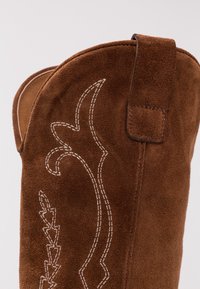 Brun ruskind cowboystøvle med indviklede hvide sy-mønstre, et fremtrædende buet design øverst og en lille løkke bagpå.