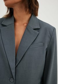 Blazer grigio realizzato in tessuto testurizzato, con rever a scollo a V, chiusura con un singolo bottone e vestibilità slim con dettaglio della tasca sul petto.