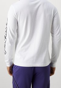 Valkoinen pitkähihaiset puuvillapaita, jossa on graafinen kuvio vasemmassa hihaosassa. Paita on yhdistetty violetin verkkomateriaalin shortseihin.