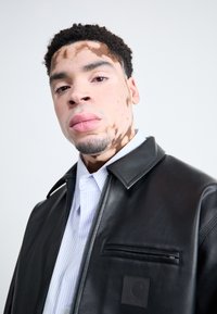 Veste en cuir noire avec une poche zippée et un logo carré, superposée à une chemise rayée bleu clair. Texture lisse, design structuré.
