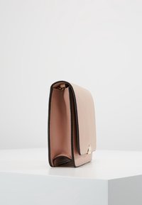 Kompakt handväska med en slät, ljusrosa läderexteriör och svart kant. Anmärkningsvärd guldfärgad detalj på framsidan.