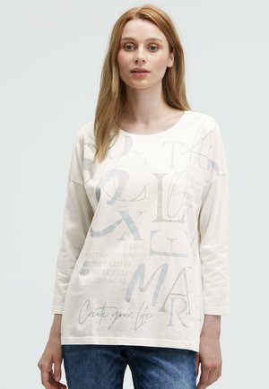 Cream long-sleeve t-shirt featuring text in gray and white, with a round neckline and relaxed fit, paired with denim jeans.
