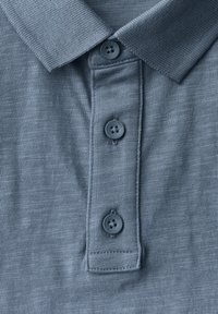 Blå polo skjorta med texturerad tyg, tre svarta knappar på knäppningen och en mjuk krage. Har ett subtilt horisontellt mönster.