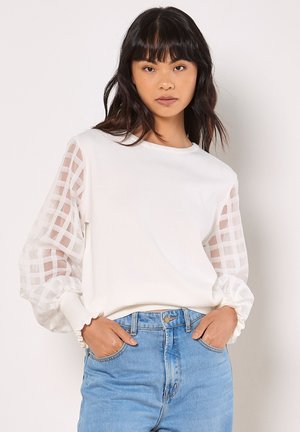 Weißer Pullover mit transparenten, karierter Musterärmeln, gerippten Bündchen und rundem Halsausschnitt, kombiniert mit hellblauen Jeans.
