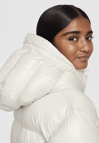 Chaqueta acolchada en blanco roto, con una gran capucha, patrón de acolchado con secciones acolchadas y una textura suave y brillante.