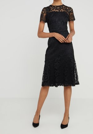 Kvinde iført knælængde sort blondekjole med korte ærmer og matchende sorte hæle, stående mod en ensfarvet lys baggrund.