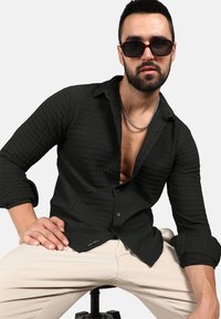 Camicia a bottone a scacchi nera con tessuto texturizzato, maniche arrotolate e scollatura a V profonda. Abbinata a pantaloni chiari.