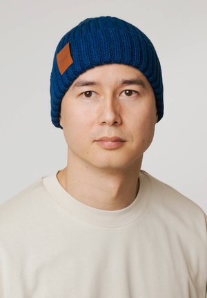 Niebieska dziana czapka z prążkowaną fakturą, z brązową skórzaną metką. Noszona przez modela w jasnej bluzie na neutralnym tle.