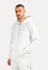 Felpa bianca con zip realizzata in tessuto morbido, dotata di tasca frontale, polsini a coste e cappuccio con cordino. Piccolo logo sul petto.