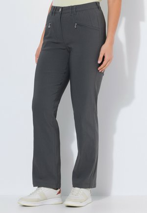 Person som bär mörkgrå raka byxor med blixtlåsfickor framtill och vita sneakers med ljusbeige och bruna detaljer.