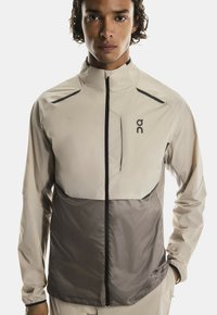 Jeune homme portant une veste d'athlétisme zippée beige et grise avec un col haut, se tenant contre un fond blanc uni.