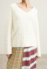 Pull en tricot blanc avec un col en V, présentant un motif texturé de triangles en relief. Porté sur une jupe plissée multicolore à rayures.