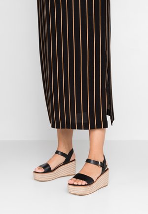 Czarne sandały na platformie z plecionym, jutowym klinem, z szerokim paskiem i klamrą z tyłu kostki, zestawione z sukienką w pionowe paski.