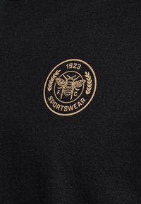 Svarta sportkläder med ett runt emblem som har en bi, lagerblad och "1923 F.C. SPORTSWEAR" i guld. Slät textur, enkel design.