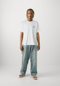 Λευκό βαμβακερό t-shirt με μικρή μπλε γραφική εικόνα στην περιοχή του στήθους. Φορεμένο με ανοιχτό μπλε φαρδύ τζιν και καφέ παπούτσια με λευκές κορδόνες.