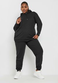 Svart ribbad huvtröja och matchande joggers, tillverkade av mjukt tyg. Innehåller en huva, långa ärmar och elastiska mudd, parat med vita sneakers.