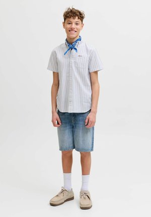 Göndör hajú tinédzser mosolyog, csíkos rövid ujjú inget, farmer rövidnadrágot, fehér zoknit, bézs cipőt és kék nyakbavalót visel.