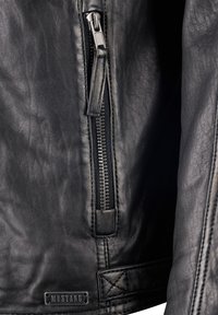 Nahaufnahme einer schwarzen Lederjacke mit vertikalem Reißverschluss mit Lederzuglasche und einem Metall-"MUSTANG"-Markenlabel, das nahe dem unteren Saum angenäht ist.
