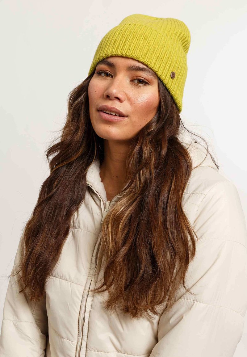 Gorro de punto amarillo con canalé y ajuste ceñido, que presenta una pequeña etiqueta de logo marrón. Llevado con una chaqueta acolchada de color beige claro.