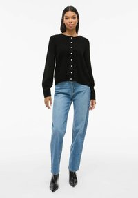 Gilet en tricot noir avec un col rond, agrémenté de boutons blancs. Associé à un jean bleu clair et des bottines noires à bout pointu.