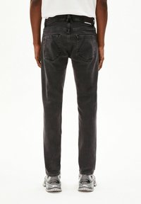 Persoon staand, gekleed in verweerde zwarte spijkerbroek met achterzakken en een wit shirt, gecombineerd met zilveren en grijze sneakers op een witte achtergrond.