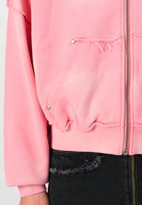 Lyserød cropped hoodie med lynlås, frynsede kanter og sølvknapdetaljer. Tekstureret stof med en afslappet pasform, parret med sorte slidte jeans.
