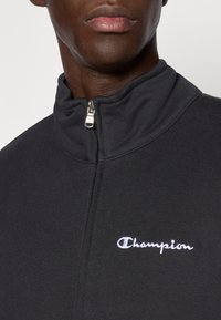 Μαύρο φούτερ με φερμουάρ και ψηλό γιακά, με λευκό κέντημα λογότυπου "Champion" στην αριστερή πλευρά. Μαλακό, υφασμένο υλικό.