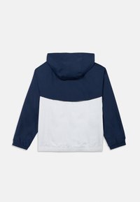 Mornarsko modra in bela vetrovka z kapuco, elastičnimi manšetami in nastavljivim spodnjim robom. Gladek občutek s kontrastnim barvnim dizajnom.
