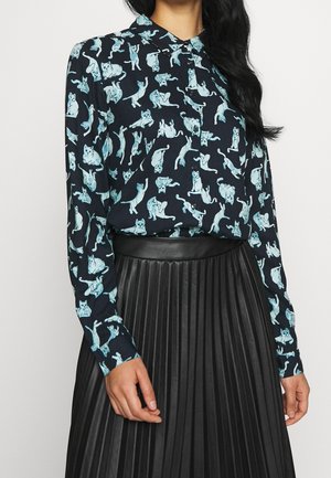 Mujer con una blusa azul marino con ilustraciones de gatos en azul claro y una falda negra plisada de talle alto.