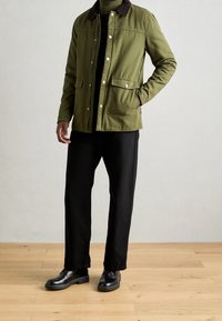 Veste vert olive avec un col marron, poches avant et boutons-pression, portée sur un haut noir et associée à un pantalon et des chaussures noirs.