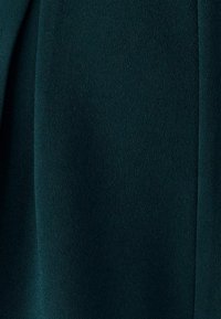 Zaļa auduma ar maigu, gludu tekstūru; raksturīgas plūstošas krokojumi un viegla spīduma mirdzums. Nav redzamas šuves vai aparatūras detaļas.