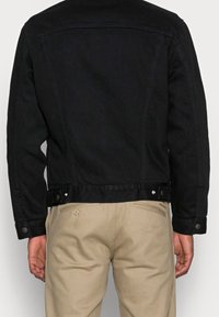 Schwarze Jeansjacke mit taillierter Passform. Mit Druckknöpfen an der Taille und einer glatten Textur. Getragen über hellbeigen Hosen.