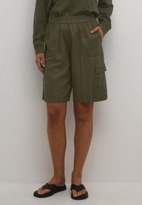 Personne portant un short cargo vert olive arrivant aux genoux avec une taille élastique, associé à des sandales noires à enfiler.