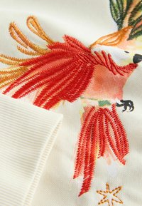 Makro záber na oranžovo-červeného vyšívaného vtáka s čiernymi pazúrmi na krémovom látke s žliabkovaným okrajom a malým vyšivaným detailom hviezdy.