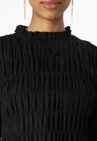 Black smocked top featuring a high ruffled neckline and textured fabric. Details include horizontal pleats and smooth, shiny earrings.