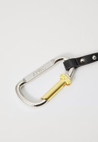 Silberner Metallkarabiner mit eingraviertem Markennamen, der an einem schwarzen Lederriemen mit goldenen und strukturierten Akzenten befestigt ist.