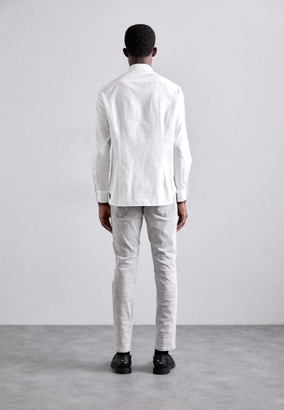 White long-sleeve shirt with a textured finish, paired with light gray slim-fit pants and black shoes, viewed from the back.