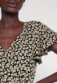 Musta mekko, jossa on V-pääntie, lyhyet röyhelöhihat ja beige kukkakuosi. Malli esittelee kankaan tekstuurin ja väljähkön istuvuuden.