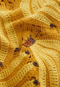 Nærbillede af gul og sennepsfarvet hæklet stof med teksturerede striber og små brune og rustfarvede strikkede detaljer.