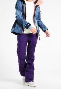 Giacca impermeabile con motivo blu e navy, dettagli in rosa, abbinata a pantaloni impermeabili viola. Dotata di cappuccio e chiusure con zip.