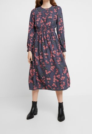 Frau trägt ein graues Midikleid mit pinkem Blumenmuster, elastischer Taille, langen Ärmeln und schwarzen Stiefeletten, steht auf einem weißen Boden.