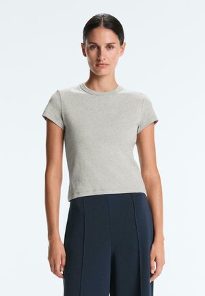 Frau mit zurückgebundenem dunklem Haar, die ein eng anliegendes hellgraues Kurzarmshirt und hochtaillierte dunkelblaue Hosen trägt, stehend vor einem schlichten Hintergrund.