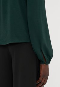 Dark green blouse with a relaxed shape, long puffed sleeves, and a side slit at the cuffs, featuring a small gold accent button. Black pants.