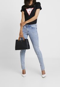 Czarna torebka na ramię z brązowymi uchwytami, o sztywnym kształcie i subtelnym brandingu. Założona z czarnym t-shirtem z czerwonym trójkątnym logo i jasnoniebieskimi dżinsami.