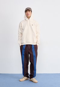 Hoodie bege com bolso canguru, calças de fato castanhas com riscas azuis, e sapatilhas bege, sobre um fundo simples.