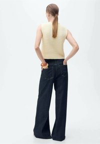 Dzianinowy bezrękawnik w jasnym kremowym kolorze zestawiony z wysokim stanem, szerokimi nogawkami i ciemnymi dżinsami z pięcioma klasycznymi kieszeniami.