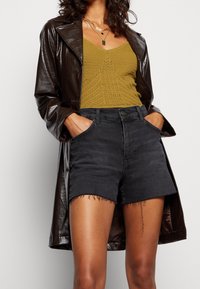 Žena v černých šortkách s vysokým pasem a roztrhanými okraji, hořčicovém pleteném topu, tmavě hnědém kabátu z umělé kůže a s vrstvami zlatých náhrdelníků.